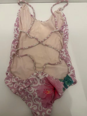 Rosa baddräkt med mönster och blomma - Säljer en unik baddräkt i ljusrosa med vita snirkliga mönster och en stor rosa blomma framtill. Baddräkten har smala axelband och coola utskärningar på sidorna. Materialet är troligtvis syntetiskt, perfekt för stranden eller poolen.