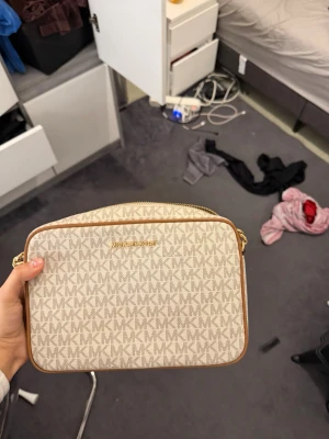 Beige Michael Kors handväska - Snygg beige handväska från Michael Kors med MK-logga över hela väskan. Väskan har guldiga dragkedjor och detaljer samt justerbar axelrem. Tillverkad i canvas med läderdetaljer och har ett ljust innerfoder med logotyp. Perfekt för dig som gillar stilrena accessoarer.äkta från Zalando köpt för 3250