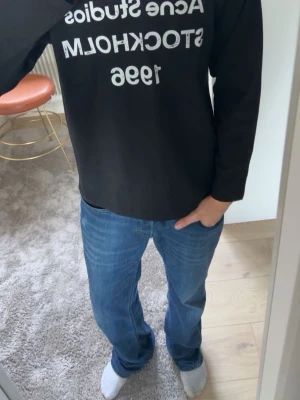 Svart långärmad topp Acne Studios - Svart långärmad topp från Acne Studios med vit textprint framtill där det står 'Acne Studios Stockholm 1996'. Klassisk passform och rund halsringning. Perfekt till jeans eller kjol för en clean och stilren look.