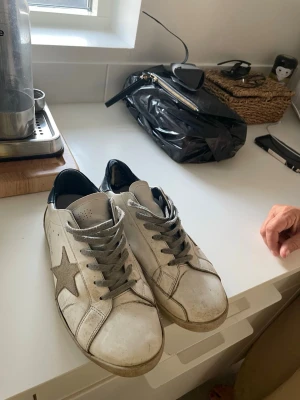 Vita Golden Goose sneakers med stjärna - Säljer ett par vita Golden Goose sneakers med grå stjärna på sidan, grå skosnören och svart detalj vid hälen. Skorna har en klassisk låg profil och är tillverkade i läder med slitna detaljer för en cool, avslappnad look. Ganska slitna och kommer även fräscha upp skorna vid köp!