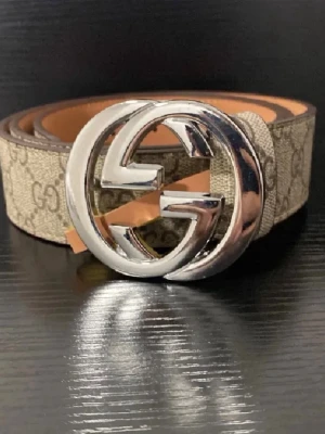 Beigebälte - Snyggt beige bälte från med klassiskoch stor silverfärgad logga som spänne. Bältet har bruna detaljer och är tillverkat i canvas med skinn på insidan. Perfekt accessoar för att lyfta din outfit.