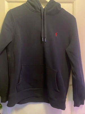 Marinblå hoodie från Polo Ralph Lauren - Snygg marinblå hoodie från Polo Ralph Lauren med klassisk röd logga broderad på bröstet. Tröjan har huva med dragsko, stor magficka och ribbade muddar vid ärmslut och nederkant. Perfekt för en avslappnad och stilren look.
