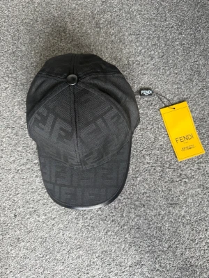 Svart keps från Fendi med FF-mönster - Snygg svart keps från Fendi med det klassiska FF-logomönstret över hela kepsen. Kepsen har böjd skärm och justerbar rem bak. Tillverkad i ett slitstarkt tyg och levereras med original Fendi-tag. Perfekt för dig som vill ha en exklusiv och trendig accessoar.