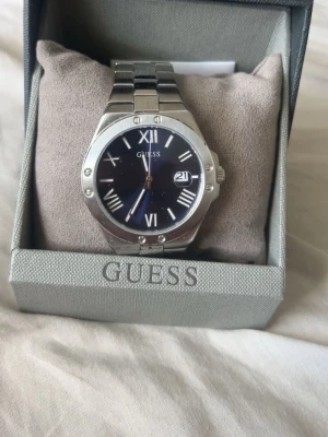 Silverfärgad klocka från Guess - Stilren klocka från Guess med silverfärgat armband i metall och mörkblå urtavla. Klockan har romerska siffror, datumvisning och robust boett. Perfekt accessoar för dig som vill ha en klassisk men modern look. Äkta 