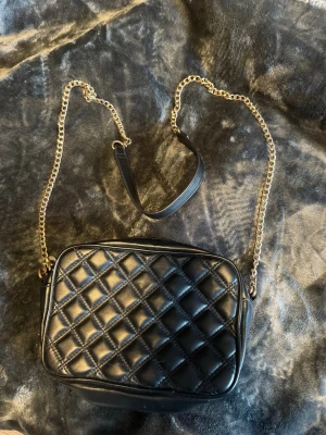 Svart axelväska med guldkedja - Snygg svart axelväska med guldfärgad kedjerem och detaljer i läderimitation. Väskan har en stilren, rektangulär form och passar perfekt för dig som gillar en trendig och elegant look. Enkel att matcha med olika outfits och ger en lyxig känsla.