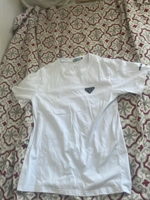 Vit t-shirt från Prada med logga - Säljer en stilren vit t-shirt från Prada med den ikoniska metalloggan på bröstet och diskret tryck på ärmen. Klassisk passform och rund halsringning. Perfekt basplagg för dig som gillar exklusiva märken och enkel design.