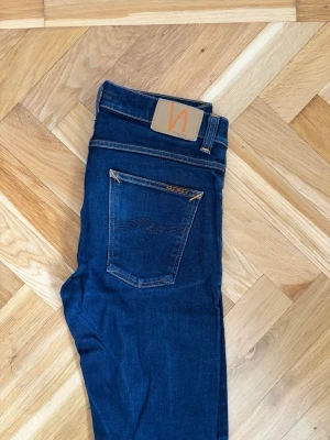 Mörkblå jeans från Nudie - Säljer ett par mörkblå skinny jeans från Nudie med klassiska orange sömmar och femficksmodell. Jeansen har hög midja och är tillverkade i stretchigt denim för en bekväm passform. Nudie-logga på bakfickan och patch i läder baktill.