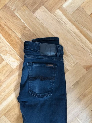 Nudie jeans  - Säljer ett par  jeans från Nudie Jeans med klassisk femficksdesign och svarta detaljer på bakfickan. Jeansen har en smal passform och är tillverkade i stretchigt denimtyg för extra komfort.