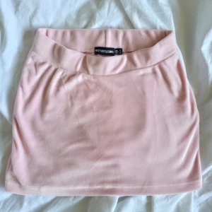Ljusrosa sammetskjol PrettyLittleThing - Supersöt ljusrosa kortkjol från PrettyLittleThing i mjuk sammet. Kjolen har en enkel, tight passform och bred linning som ger en clean och trendig look. Perfekt att styla med crop top eller oversized hoodie. Passar storlek XS och S.