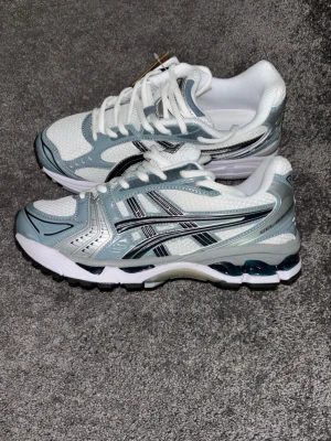 asics gel kayano 14 - Snygga sneakers från Asics i vitt och ljusgrått med svarta detaljer. Skorna har en sportig design med mesh- och syntetmaterial, samt klassiska Asics-stripes på sidorna. Perfekta för dig som gillar streetwear och bekväma skor.