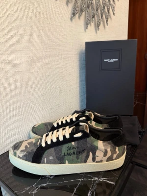 Saint Laurent camo sneakers - Snygga sneakers från Saint Laurent med camouflagemönster i grönt, beige och svart. Skorna har vita snören, svart mockadetalj vid hälen och vit sula. Märkeslogga broderad på sidan. Perfekta för dig som vill sticka ut med en cool streetstyle.