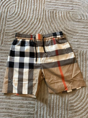 Burberry shorts - Säljer ett par klassiska Burberry shorts med rutigt mönster i beige, svart, vit och röd. Shortsen har elastisk midja med svart snörning och en bakficka. Perfekta för sommaren och riktigt snygga till streetwear-stilen. Storlek XL fast de är småa i storlek och passar L och M. Oanvända och helt nya så pris kan diskuteras!