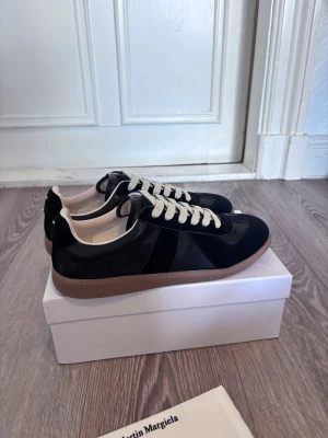 Svarta sneakers från Maison Margiela - Svarta sneakers från Maison Margiela med beige sula och vita skosnören. Ovandelen är i en mix av mocka och läder, vilket ger en clean och stilren look. Klassisk låg modell som passar till det mesta och har diskreta detaljer för en exklusiv känsla.