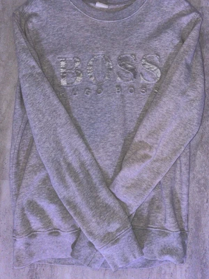 Grå sweatshirt från Hugo Boss - Snygg grå sweatshirt från Hugo Boss med stor, glansig BOSS-logga framtill. Tröjan har rund halsringning, långa ärmar och ribbade muddar vid ärmslut och nederkant. Tillverkad i mjukt bomullsmaterial som känns skönt mot huden.
