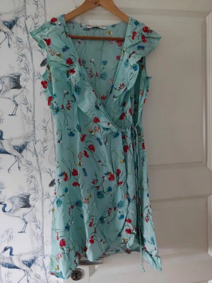 Blommig omlottklänning LCW Casual - Söt ljusblå omlottklänning från LCW Casual med blommigt mönster i rött, blått och gult. Klänningen har volangdetaljer vid ärmar och halsringning samt knytband i midjan. Perfekt för varma dagar och ger en fräsch sommarkänsla.