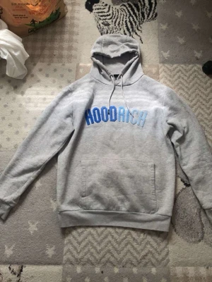 Grå hoodie från Hoodrich med tryck - Snygg grå hoodie från Hoodrich med blått tryck på bröstet. Klassisk känguruficka framtill och justerbar huva med dragsko. Perfekt för en chill och street-inspirerad look. Tillverkad i mjukt sweatshirtmaterial för extra komfort.