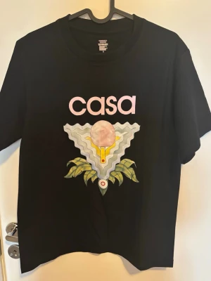 Svart t-shirt med grafiskt tryck Casa - Svart t-shirt från Casa med ett stort grafiskt tryck framtill i pastellfärger och texten 'casa' i rosa. T-shirten har rund hals och korta ärmar. Perfekt för dig som gillar unika prints och streetwear vibes.