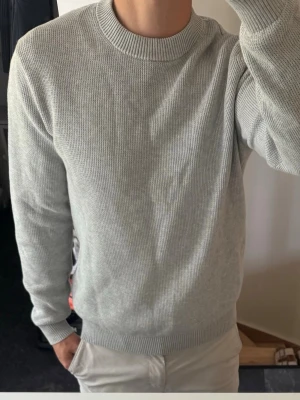 Ljusgrå stickad tröja Selected Homme - En stilren ljusgrå stickad tröja från Selected Homme med rund halsringning och ribbade muddar vid ärmslut och nederkant. Tröjan har en klassisk passform och är perfekt för lager-på-lager under kyligare dagar.
