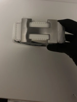 Vitt skärp med H-spänne - Snyggt vitt skärp med ett stort, silverfärgat H-format metallspänne. Skärpet har tydliga sömmar längs kanterna och är gjort i slätt skinn. Perfekt för att lyfta vilken outfit som helst.