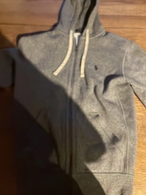 Grå zip hoodie från Polo Ralph Lauren - Snygg grå hoodie med dragkedja från Polo Ralph Lauren. Klassisk design med vit dragsko i huvan och broderad logga på bröstet. Mjuk bomullsmix och fickor framtill gör den både chill och stilren.