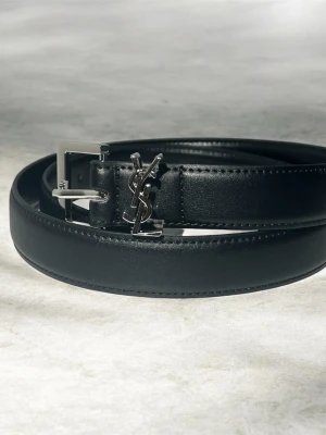 Svart bälte från Saint Laurent - Snyggt svart bälte från Saint Laurent med klassisk silverfärgad YSL-logga och spänne. Bältet är gjort i slätt skinn och har en stilren, minimalistisk design som passar till det mesta. Perfekt accessoar för att lyfta din outfit.