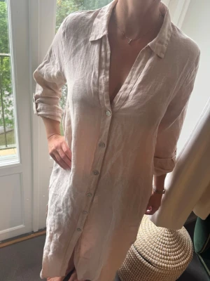 Beige linneskjortklänning med knappar - Luftig och stilren skjortklänning i beige linne med klassisk krage och knappar hela vägen framtill. Klänningen har långärmade ärmar som kan vikas upp och en avslappnad passform. Perfekt för varma dagar och enkel att styla. (Pris kan diskuteras)