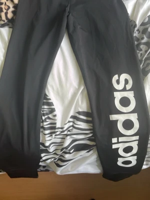 Svarta leggings från Adidas - Säljer ett par svarta leggings från Adidas med stor vit logga längs ena benet och mindre logga vid vristen. Tight passform och stretchigt material som sitter skönt. Perfekta till träning eller chill. Klassisk sportig look med hög midja.