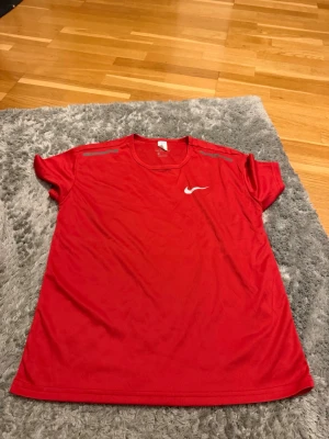 Röd Nike sporttröja - Snygg röd sporttröja från Nike med korta ärmar och rund hals. Tröjan har ett vitt Nike-logotyptryck på bröstet och diskreta grå detaljer på axlarna. Tillverkad i ett lätt och ventilerande funktionsmaterial som passar perfekt till träning.