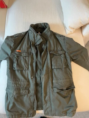 Grön Superdry field-jacket - En grön Superdry field-jacket som är väl omhändertagen och i jättebra skick. Väldigt bra kvalitet och jackan känns ”tung”. Storlek xl