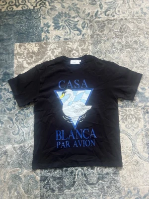 Svart Casa Blanca t-shirt med svan storlek S - Cool svart t-shirt från Casa Blanca med stor grafisk print framtill som visar en vit svan och blå triangulär bakgrund. Texten 'CASA BLANCA PAR AVION' i blått. Klassisk passform och rund hals. Perfekt för dig som gillar streetwear och unika prints. Den är i storlek S, helt ny och aldrig buren
