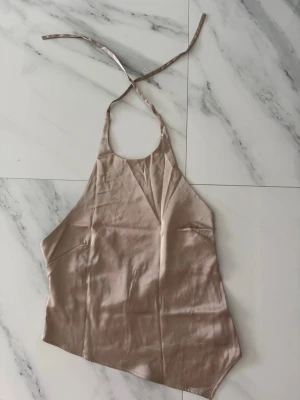 Beige satin linnetopp med knyt - Minimalistisk beige linnetopp i satin med halterneck och tunna band som knyts i nacken. Toppen är ärmlös och har en asymmetrisk nederkant för en modern vibe.