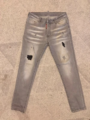 Grå slitna jeans från Dsquared2 - Säljer ett par grå skinny jeans från Dsquared2 med slitningar och hål på båda benen. Jeansen har fem fickor, dragkedjegylf och en stor röd Dsquared2-lapp bak i midjan. 
