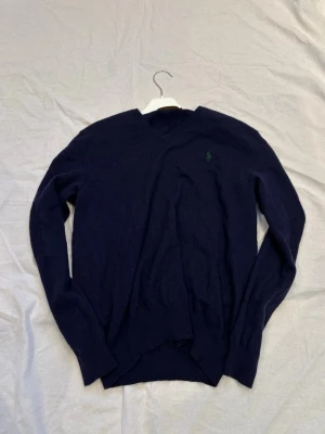 Mörkblå stickad tröja Polo Ralph Lauren - Mörkblå stickad tröja från Polo Ralph Lauren med V-ringning och den klassiska gröna logotypen broderad på bröstet. Tröjan har långa ärmar och ribbade muddar vid ärmslut och nederkant. Perfekt för en stilren och avslappnad look.