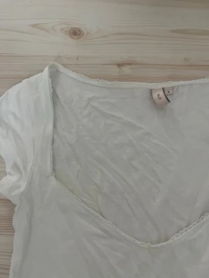 Vit v-ringad topp med spetskant - Vit topp från Nelly med djup v-ringning och diskret spetskant längs urringningen. Kortärmad och i mjukt, tunt material som känns skönt mot huden. Enkel och stilren design som passar till mycket.