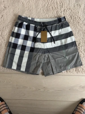 Burberry shorts - Snygga burberry badbyxor med klassiskt rutigt mönster i svart, vitt och grått. De har elastisk midja med snörning och en bakficka med samma rutiga design. Perfekta för stranden eller poolen, tillverkade i snabbtorkande material.