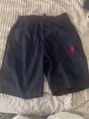 Marinblå shorts från Polo Ralph Lauren - Marinblå shorts från Polo Ralph Lauren med röd broderad logga på vänster ben. Shortsen har elastisk midja med snörning och sidofickor. Perfekta för sommardagar och avslappnad stil. Storlek S/M