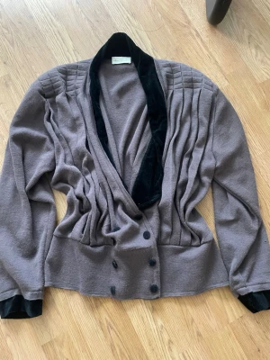 Grå vintage kofta med sammetsdetaljer - Unik brun kofta med dubbelknäppning och bred krage i svart sammet. Plisserade detaljer längs axlar och framstycke ger en cool retrovibe. Långa ärmar med matchande sammetsmuddar. Perfekt för dig som gillar statement-plagg och vintagekänsla.❤️