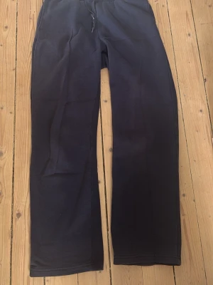 Mörkblå mjukisbyxor från Gina Tricot - Mörkblå mjukisbyxor från Gina Tricot med raka ben och dragsko i midjan. Byxorna har resår i midjan och är tillverkade i ett mjukt och bekvämt material, perfekt för chill dagar. Klassisk och enkel design utan tryck eller detaljer.