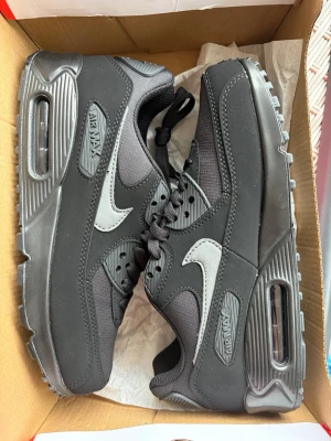 Nike Air Max 90 svarta sneakers - Snygga Nike Air Max 90 sneakers i svart med grå swoosh och detaljer. Skorna har en klassisk siluett med synlig Air Max-dämpning i sulan, robust yttersula och ovandel i en mix av läder och mesh. Perfekta för dig som gillar streetwear och sportig stil.