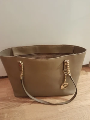 Guldbeige Michael Kors handväska - Stilren guldbeige handväska från Michael Kors med guldfärgade kedjedetaljer och MK-logga. Väskan har en klassisk rektangulär form och är tillverkad i slätt läder. Perfekt för dig som gillar snygga accessoarer med exklusiv känsla.