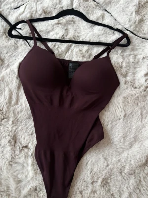 Vinröd body från H&M med vadderade kupor - Snygg vinröd body från H&M med tunna axelband och vadderade kupor. Tillverkad i ett mjukt och stretchigt material som sitter skönt mot kroppen. Perfekt att styla under en skjorta eller kavaj för en trendig look.