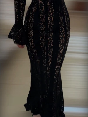 Svart lång spetsklänning - Elegant lång klänning i svart spets med transparenta detaljer och figurnära passform. Klänningen har långa ärmar med utsvängda ärmslut och ett vackert spetsmönster som ger en lyxig känsla. Perfekt för dig som vill sticka ut med en dramatisk look.