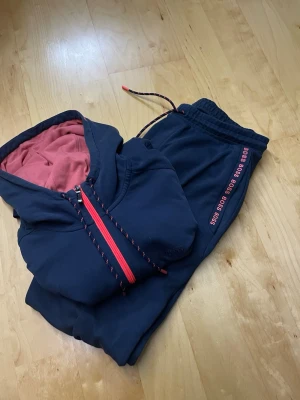 Mörkblå hoodie från BOSS med dragkedja - Säljer en mörkblå hoodie från BOSS med röd dragkedja och matchande snören. Insidan av huvan är rosa och byxorna har BOSS-logga i rött längs benet. Perfekt för en avslappnad och sportig stil.