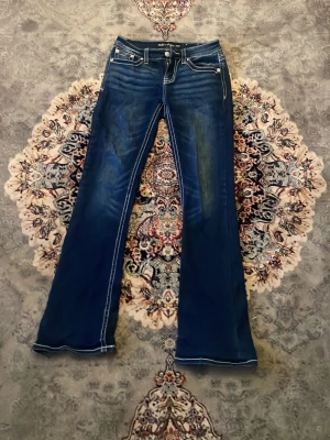 Blå bootcut jeans med broderi - Säljer ett par blå bootcut jeans med vita kontrastsömmar och snygga broderade detaljer på bakfickorna. Jeansen har låg midja och klassisk femficksmodell. Perfekta för dig som gillar en retro och unik stil.