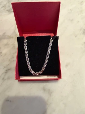 Silverfärgat tvinnat halsband - Snyggt halsband i silverfärgad metall med tvinnad kedja. Halsbandet har en klassisk karbinhake och kommer i en röd presentask. Perfekt accessoar för att lyfta din stil. Kedjan är i rostfritt stål och är inte äkta 