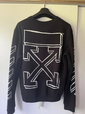 Svart sweatshirt från Off-White - Cool svart sweatshirt från Off-White med ikoniska vita pilar och grafiska detaljer på ryggen samt ränder på ärmarna. Rund halsringning och relaxed fit. Perfekt för dig som gillar streetwear och vill sticka ut med en unik design.