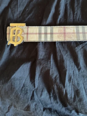  Burberry bälte med guldspänne - Säljer ett klassiskt rutig bälte från Burberry med ikoniskt guldigt TB-spänne. Bältet har beige, bruna och röda toner i det typiska Burberry-mönstret. Perfekt accessoar för att lyfta din outfit.