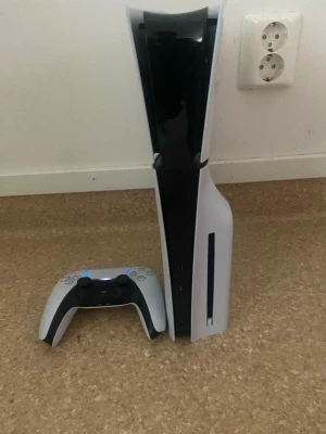PlayStation 5 med handkontroll - PlayStation 5 konsol från Sony i vit och svart design med futuristiska former. Konsolen har en diskret skåra för skivor och medföljer gör en matchande DualSense-handkontroll. Materialet är plast med matta och blanka ytor. Perfekt för gaming på nästa nivå.
