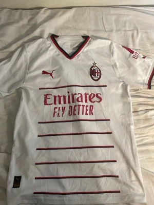 AC Milan vit matchtröja Puma - Snygg vit AC Milan matchtröja från Puma med röda detaljer och klubbmärke på bröstet. Tröjan har V-ringning med röd och svart kant, röda ränder på framsidan och sponsortryck 'Emirates Fly Better'. Tillverkad i lätt och ventilerande material.