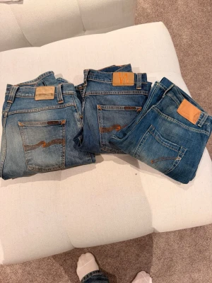 Blå jeans från Nudie Jeans - Tre par klassiska blå jeans från Nudie Jeans med bruna sömmar och läderpatch bak i midjan. Jeansen har raka ben och fem fickor. Perfekta för en avslappnad och trendig stil. Tillverkade i slitstarkt denimtyg. Olika stl mellan 31-34 